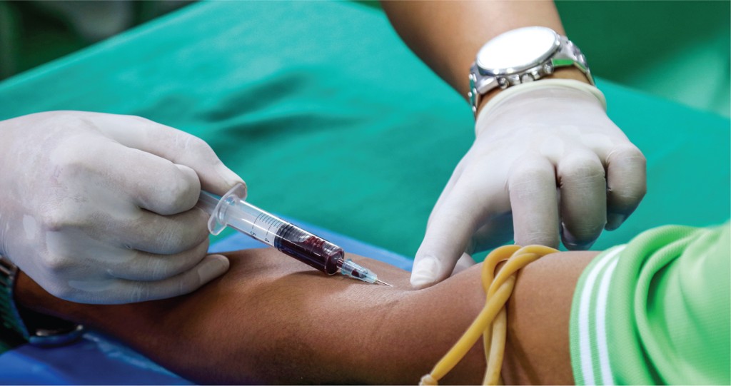 A clinician in white gloves draws blood from a patient's forearm with a syringe, a yellow tourniquet around the upper arm.