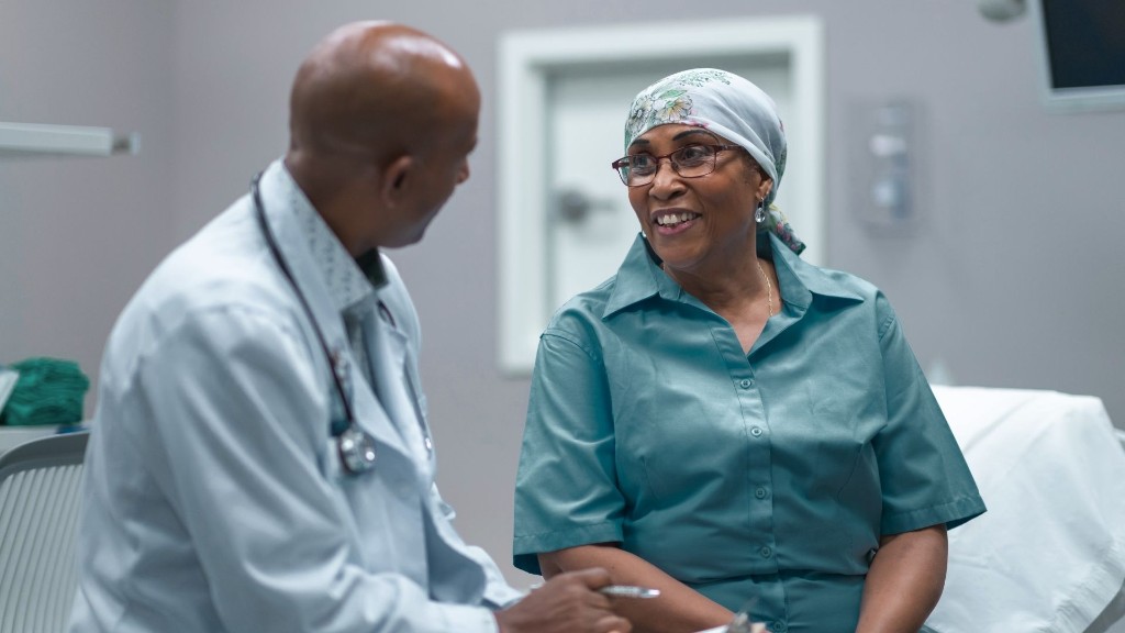 A clinician in a white coat with a stethoscope talks with a smiling patient in glasses and a headscarf during a consultation.